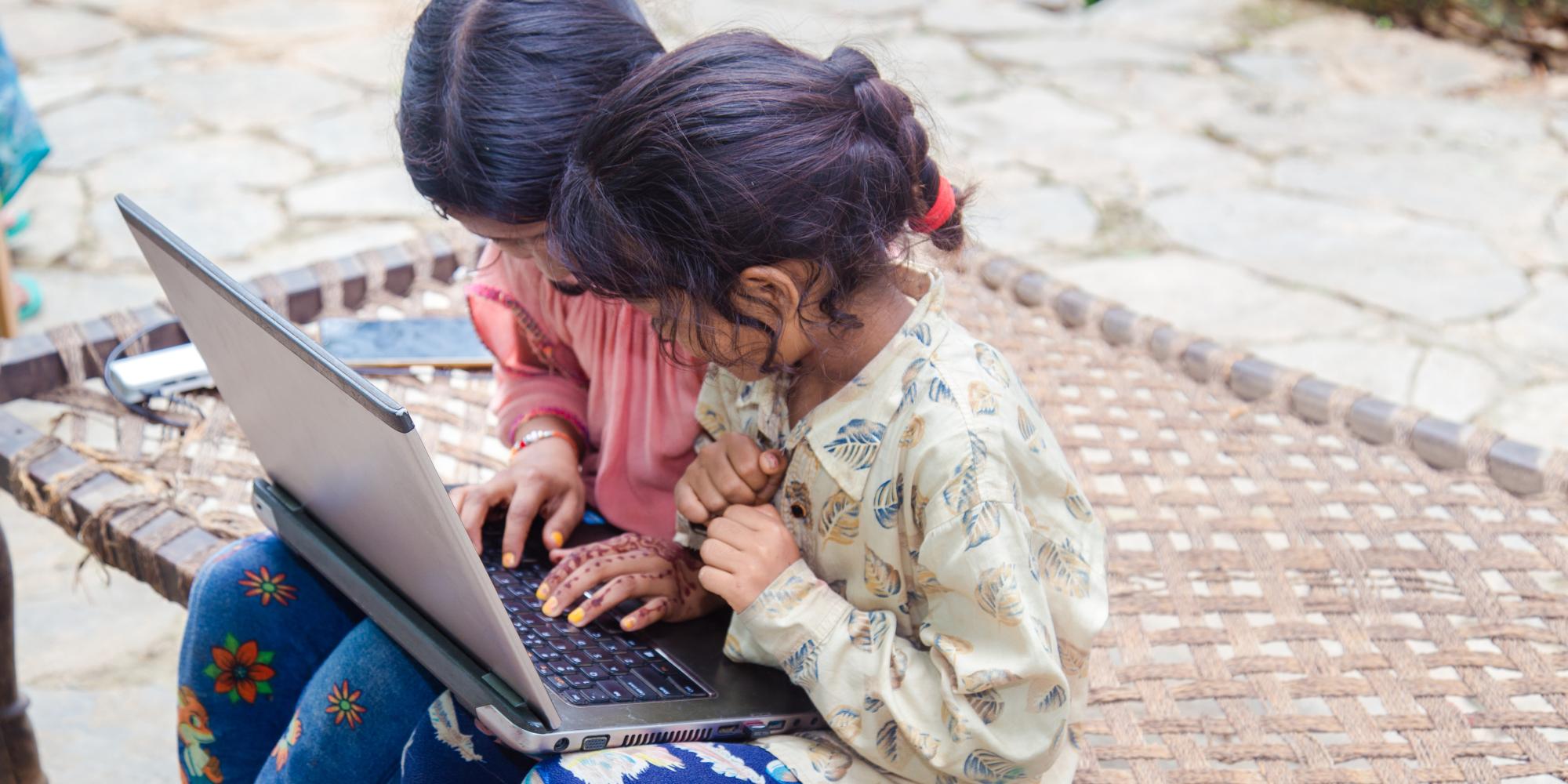 Two girls sitting on the pavement, looking at a computer.